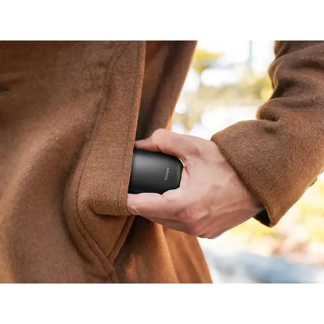 A hand puts the black Ocoopa UT3 Lite Magnetic Rechargeable Hand Warmers into a brown coat pocket outdoors.