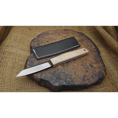 A natural maple Ohta Knives FK7 Folder rests open on a dark stone with its charcoal sheath behind it, set against a burlap background.