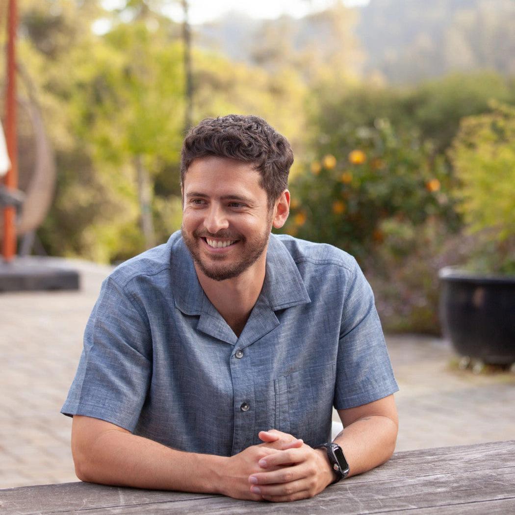A man wearing the Old Ranch Men's Bodi Shirt in Dark Blue sits at a table outdoors with trees and a potted plant in the background.