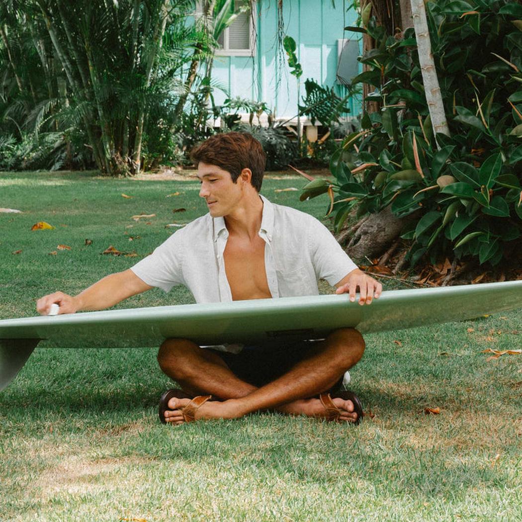 A man sits cross-legged on grass holding a surfboard, wearing the Tan Dark Java Olukai Men's Hikianalia sandals in a tropical outdoor setting.