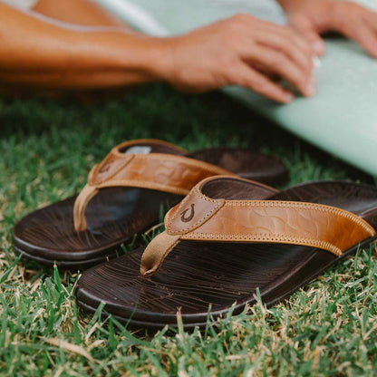 A pair of Tan Dark Java Olukai Men's Hikianalia sandals lie on green grass with a person’s hands resting on a light green board in the background.