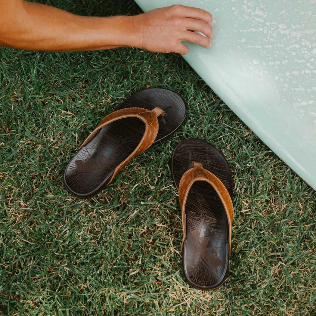 A pair of Olukai Men's Hikianalia Tan Dark Java sandals lie on green grass next to a light blue surfboard.