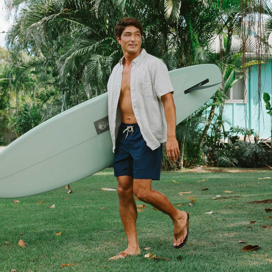 A man wearing the Tan Dark Java Olukai Men's Hikianalia sandals walks on grass with a surfboard, in front of a turquoise house and palm trees.