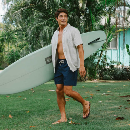 A man wearing the Tan Dark Java Olukai Men's Hikianalia sandals walks on grass with a surfboard, in front of a turquoise house and palm trees.