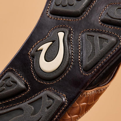A close-up of the Olukai Men's Hikianalia in Tan Dark Java showing the logo and textured sole against a neutral background.