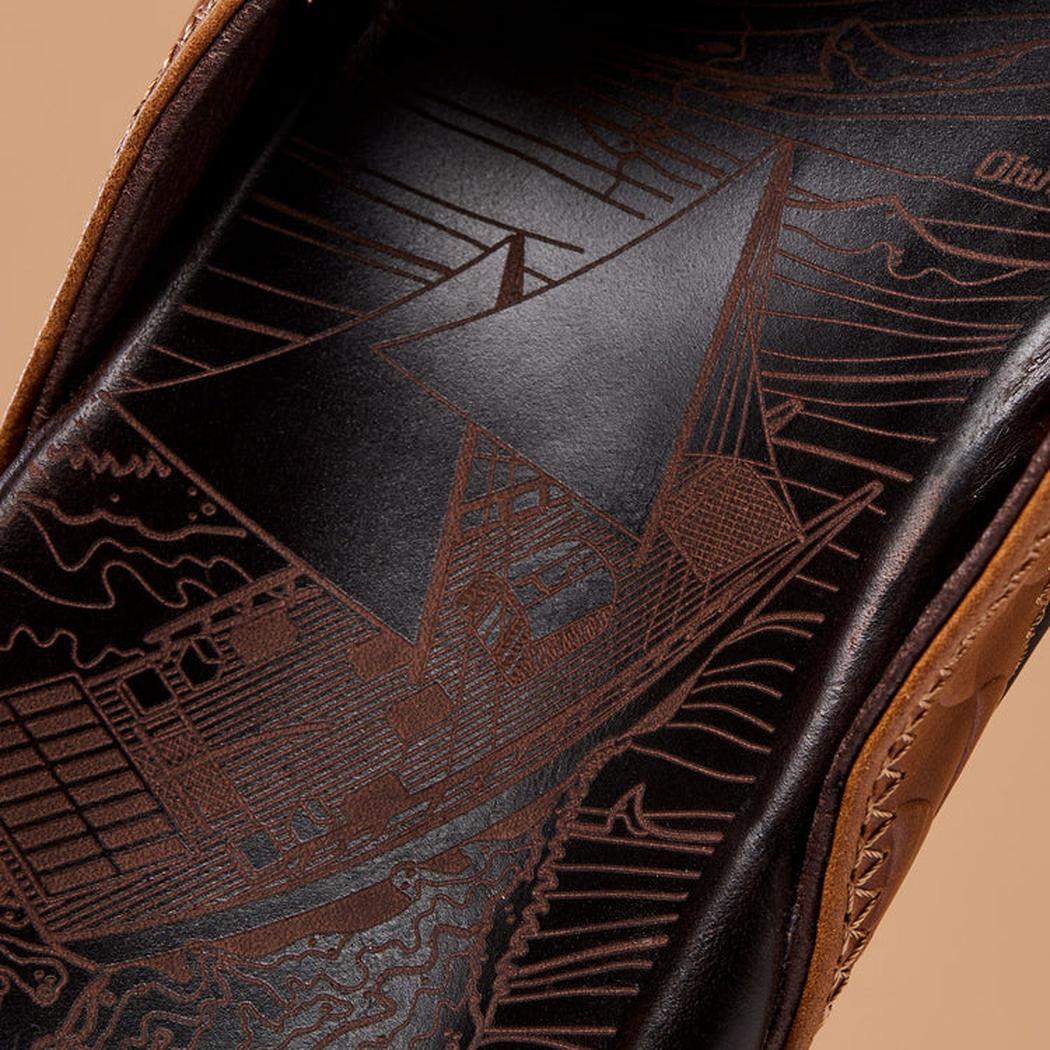 The insole of the Olukai Men's Hikianalia Tan Dark Java sandals features a detailed sailboat design against a dark background.