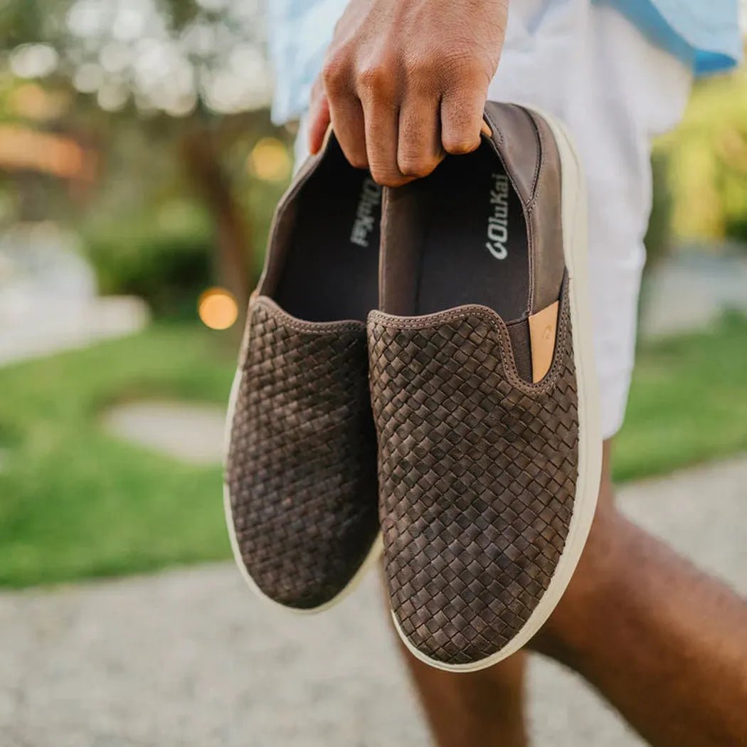 A hand holds a pair of Olukai Men's Lae'Ahi Lauhala in Dark Java, showing the woven upper and white rubber sole against a grassy outdoor background.