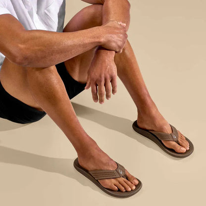 A man wearing Olukai Men's Ulele Mustang sandals sits with crossed legs on a light background.