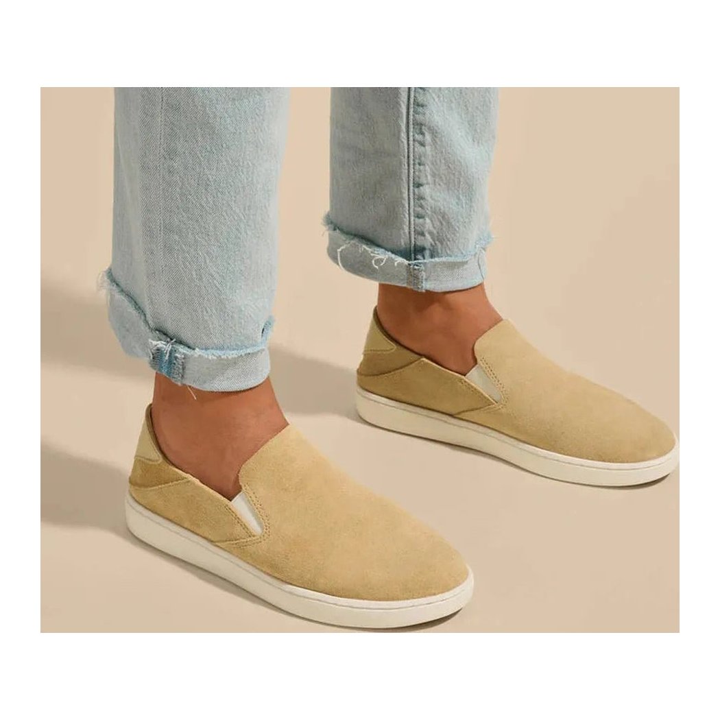 Side view of the Olukai Women's Pehuea Luxe in Toastd Coconut showing the slip-on design on a model wearing light blue jeans against a neutral background.