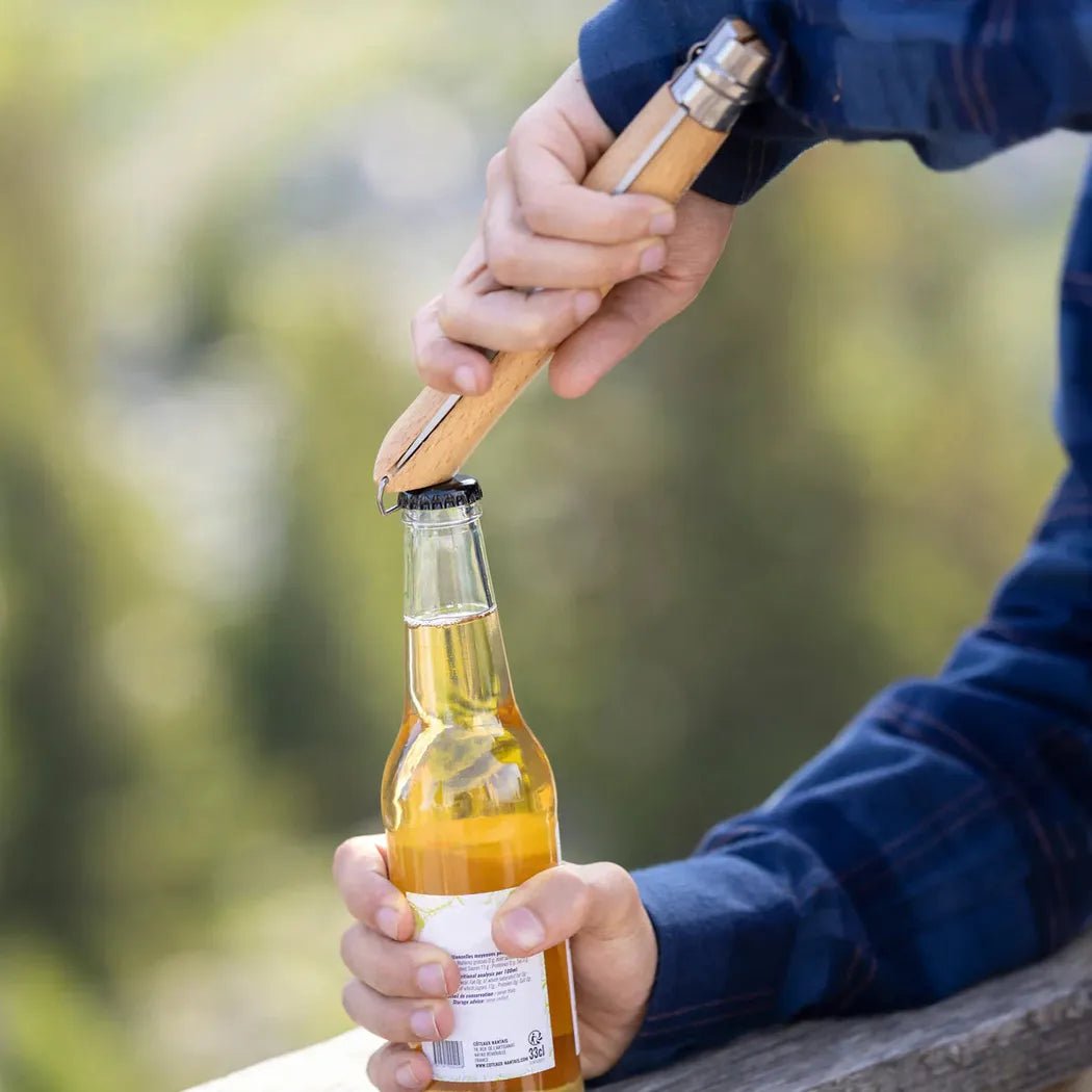 A person uses the Opinel Grilling Set to open a beer bottle while sitting outdoors on a wooden railing with green trees in the background.