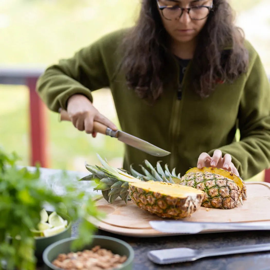 A woman in a green fleece uses a knife from the Opinel Grilling Set to cut a pineapple on an outdoor table.