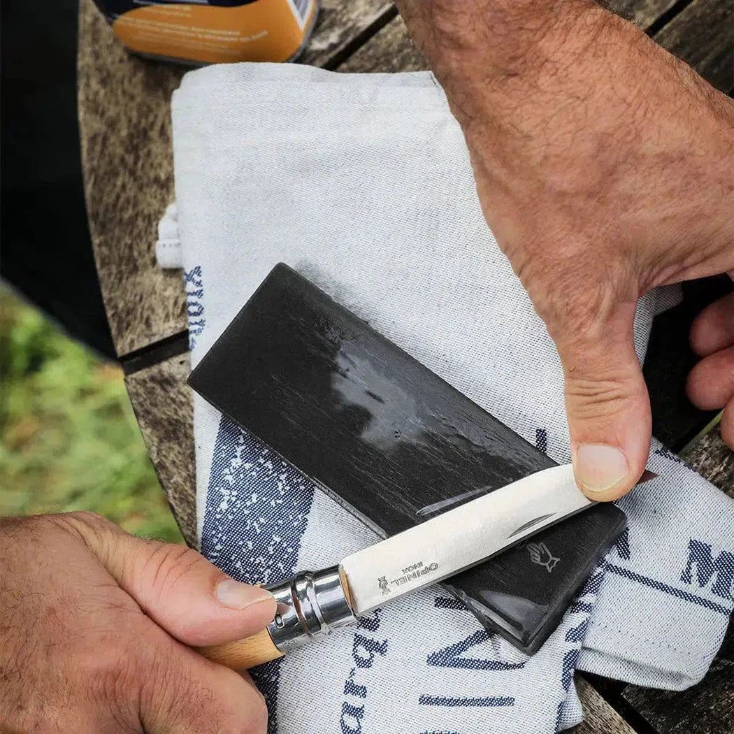 A hand holds a knife over a charcoal-colored sharpening stone on a white cloth on a wooden table.