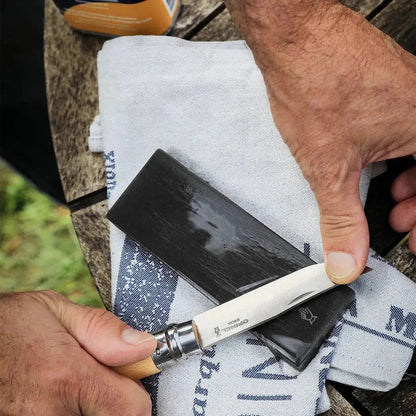 A hand holds a knife over a charcoal-colored sharpening stone on a white cloth on a wooden table.