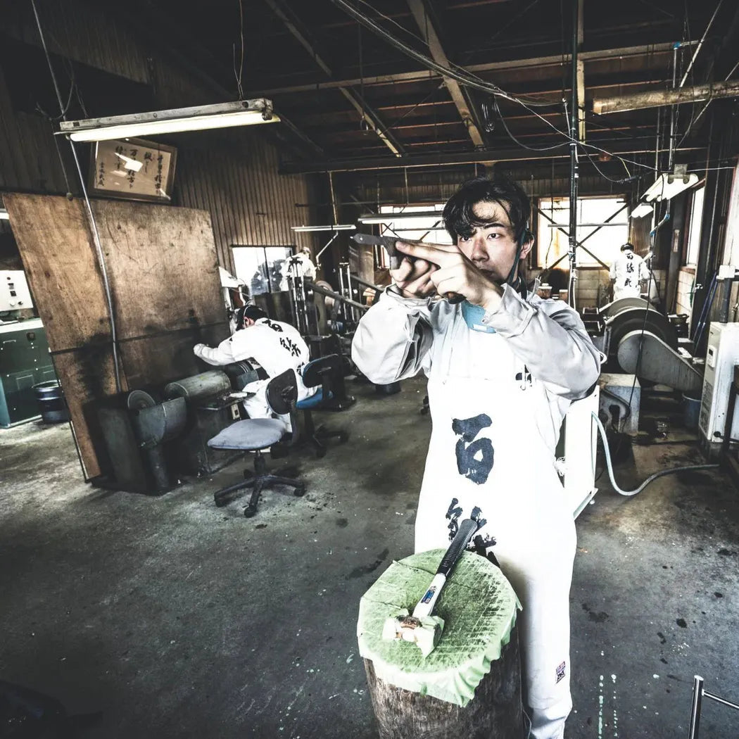 A man in a workshop wearing the Opinel Limited Edition No.10 Shiori knife points at the camera with a tree stump and knife block in front of him.