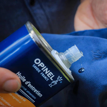 A close-up of the blue Opinel Maintenance Kit with its nozzle visible, held in a hand against a blue fabric background.