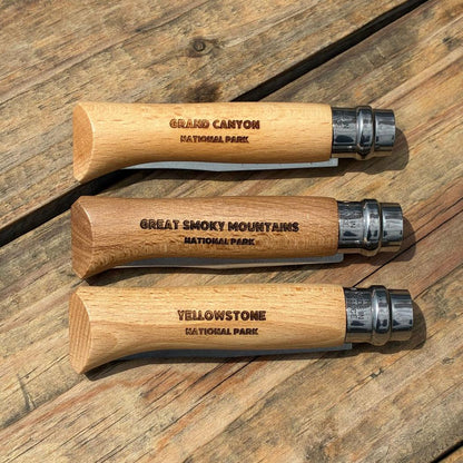 Three wooden-handled Opinel National Parks Collection knives with Grand Canyon National Park engraving lie on a wooden surface.
