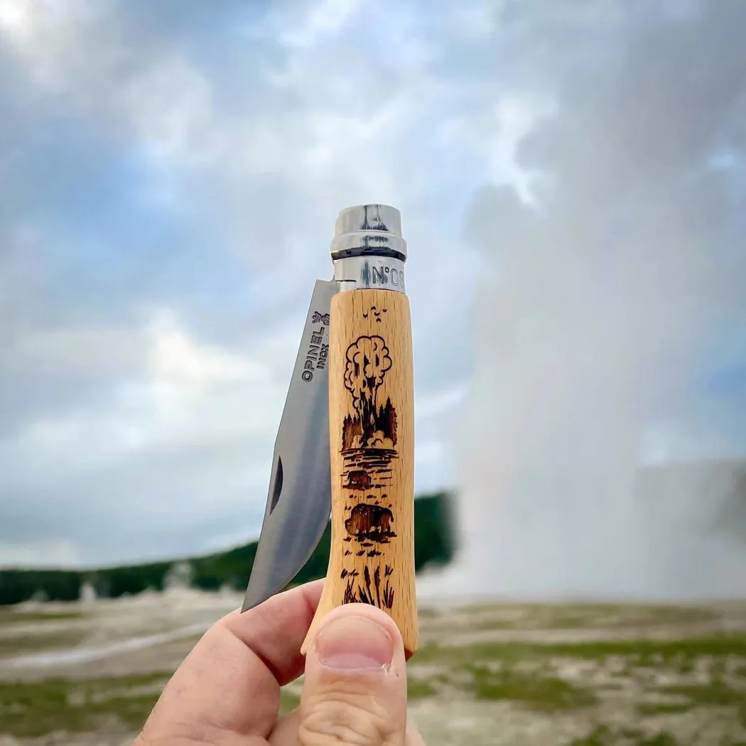 A hand holds the Opinel National Parks Collection Grand Canyon knife with a geyser in the background.