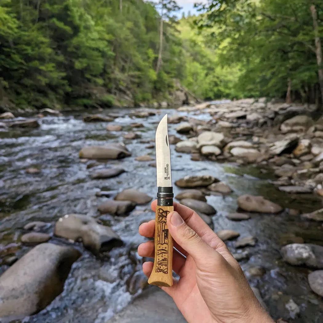 A hand holds the Opinel National Parks Collection Grand Canyon knife in front of a flowing river and forested background.