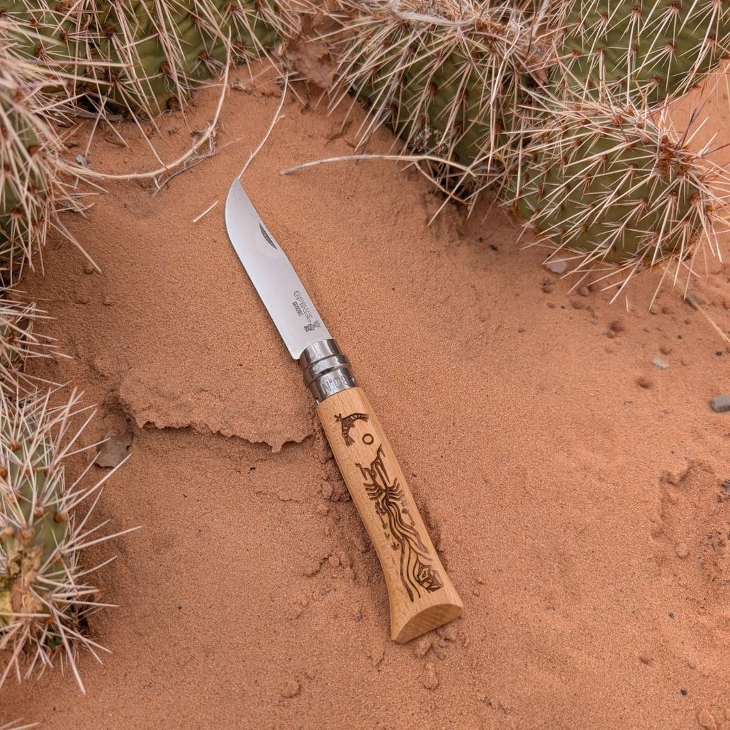 A Grand Canyon colored Opinel National Parks Collection knife lies on red sand with cactus in the background.