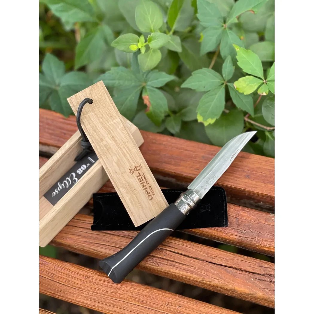 A side view of the Opinel No.08 Ellipse Ebony Folding Knife on a wooden bench with green foliage in the background.