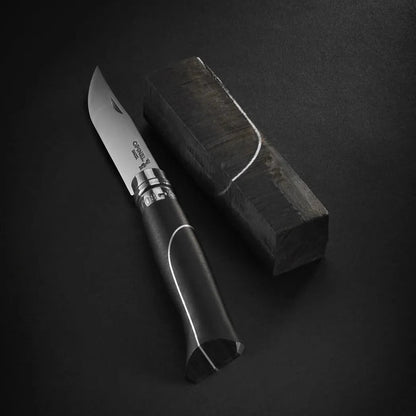 A side view of the Opinel No.08 Ellipse Ebony Folding Knife with its black handle and silver accents resting next to a charcoal-colored box on a dark background.