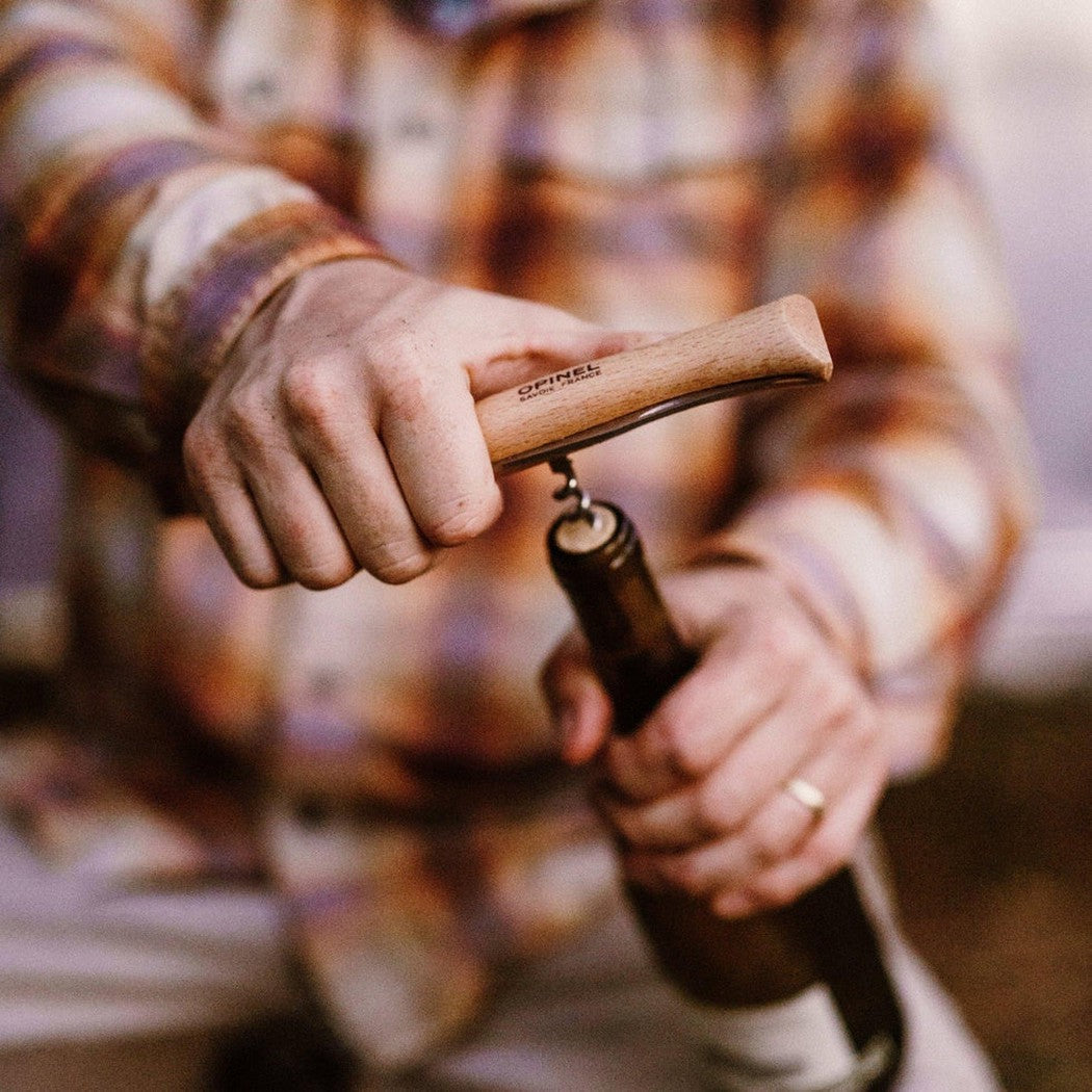 A person in a plaid shirt uses the Opinel No.10 Corkscrew Knife to open a wine bottle, with the wooden handle clearly visible.