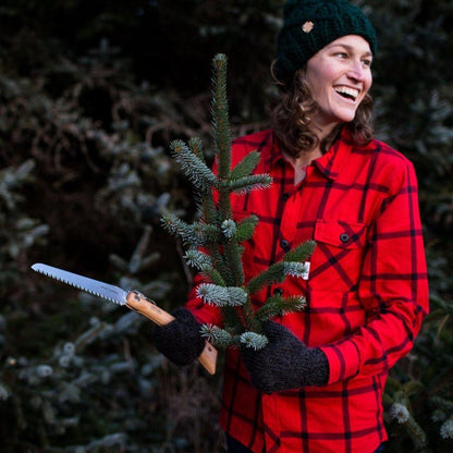 A woman in a red plaid shirt holds a small evergreen tree and the Opinel No.18 Carbon Steel Folding Garden Saw, smiling in a forested background.