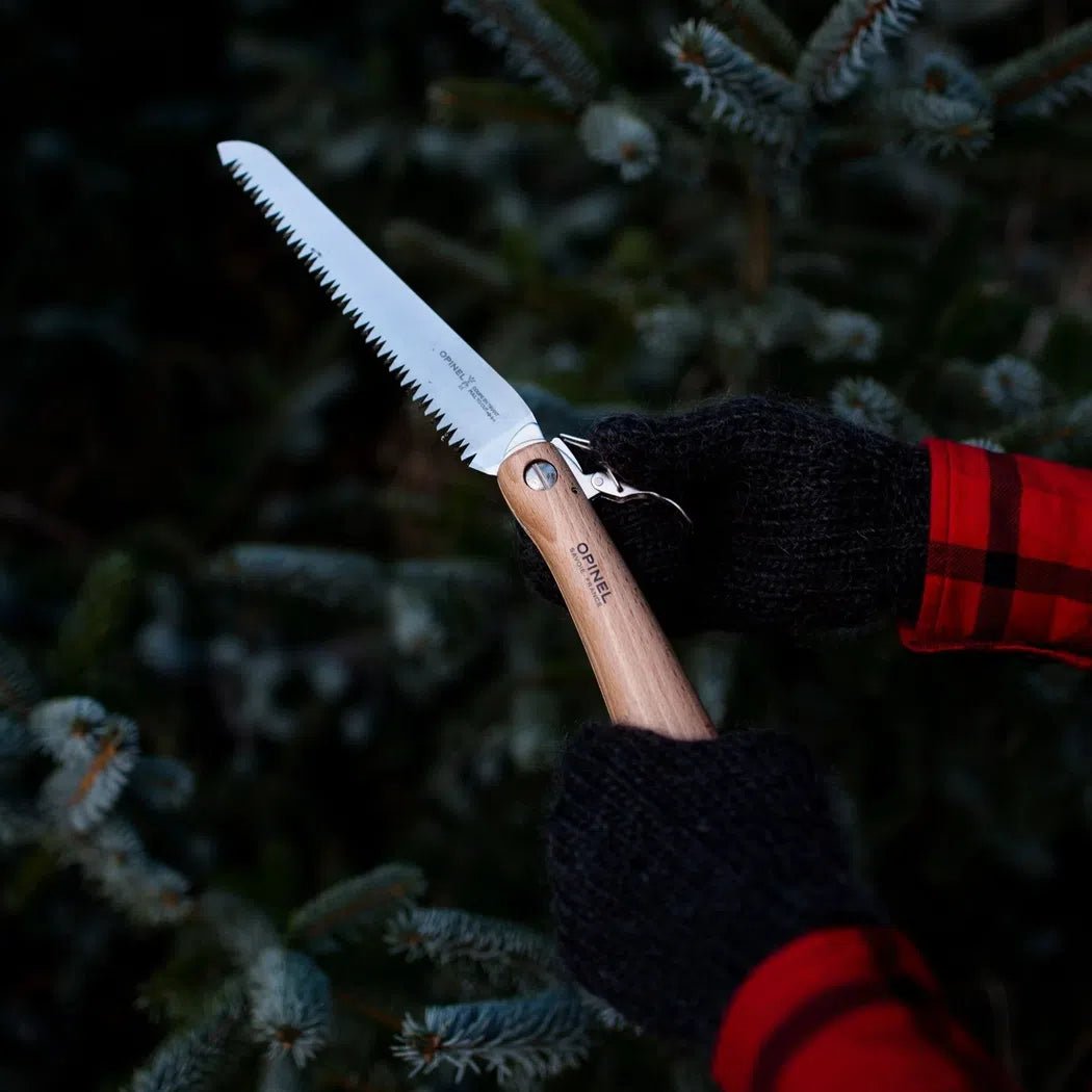 A person wearing black and red plaid gloves holds the Opinel No.18 Carbon Steel Folding Garden Saw in front of a snowy evergreen tree.