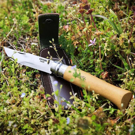 A close-up of the Opinel No.8 Stainless & Sheath knife with its wooden handle and metal bolster resting on a dark brown leather sheath in a grassy outdoor setting.