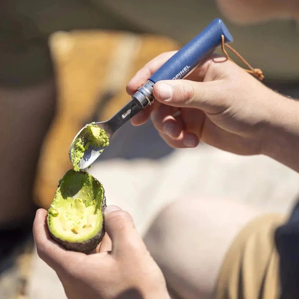A person uses the blue Opinel Picnic Plus Cutlery Insert to scoop avocado from a halved avocado on a light surface.