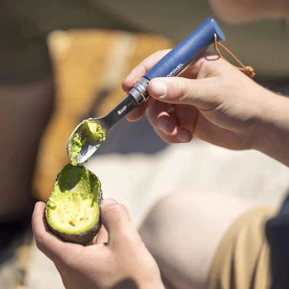 A person uses the blue Opinel Picnic Plus Cutlery Insert to scoop avocado from a halved avocado on a light surface.