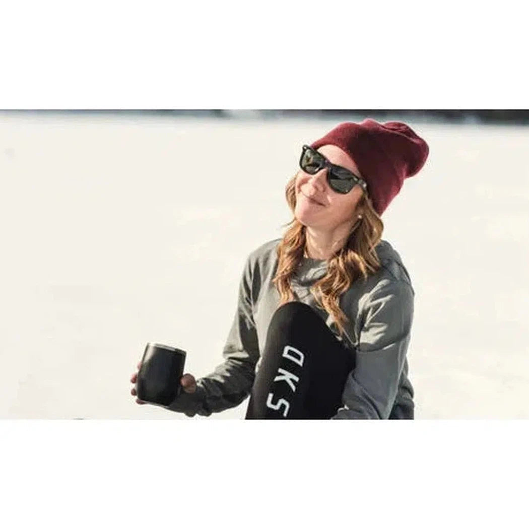 A woman in a maroon beanie and gray jacket wears Shiny Black Optic Nerve Dylan sunglasses while holding a black cup on a snowy background.