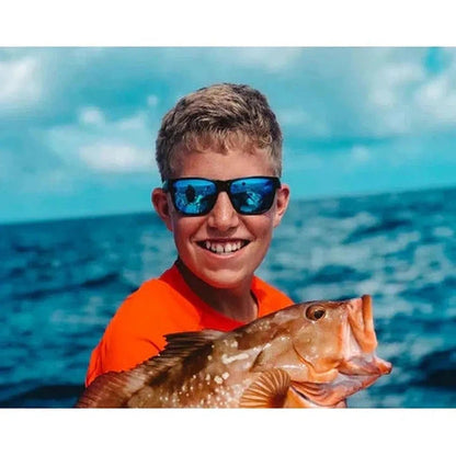 A smiling boy in an orange shirt holds a large fish while wearing the Optic Nerve Kingfish sunglasses against a blue ocean background.