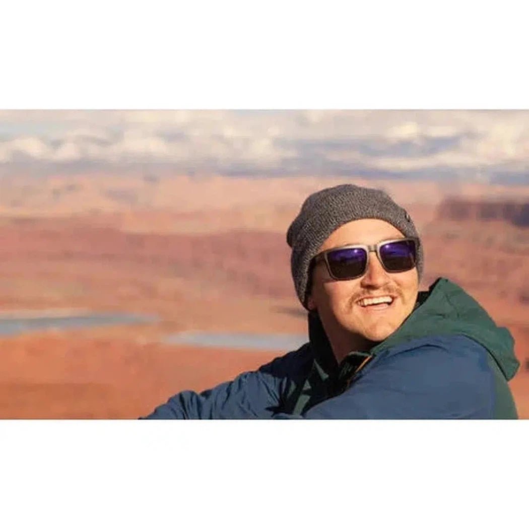 A smiling model wears the Optic Nerve Mashup sunglasses in Matte Black + Wood against a desert landscape.