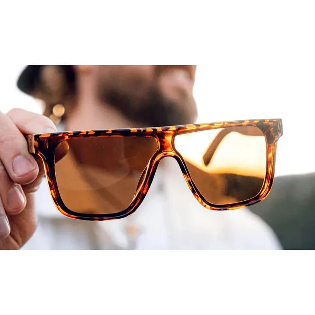 A hand holds the Shiny Black + Wood Optic Nerve Mojo sunglasses with a blurred outdoor background.