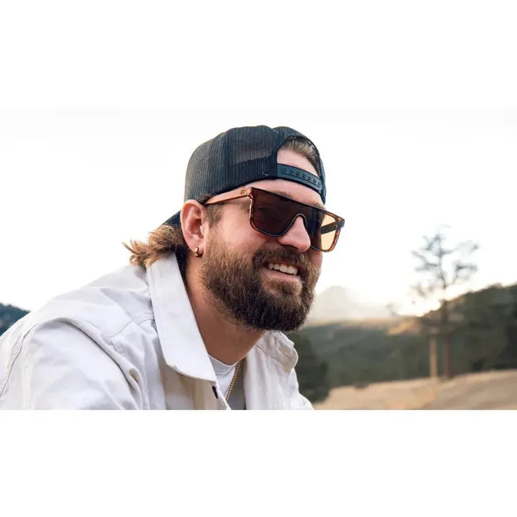 A man wearing Shiny Black + Wood Optic Nerve Mojo sunglasses smiles in a sunny outdoor setting with trees and mountains in the background.
