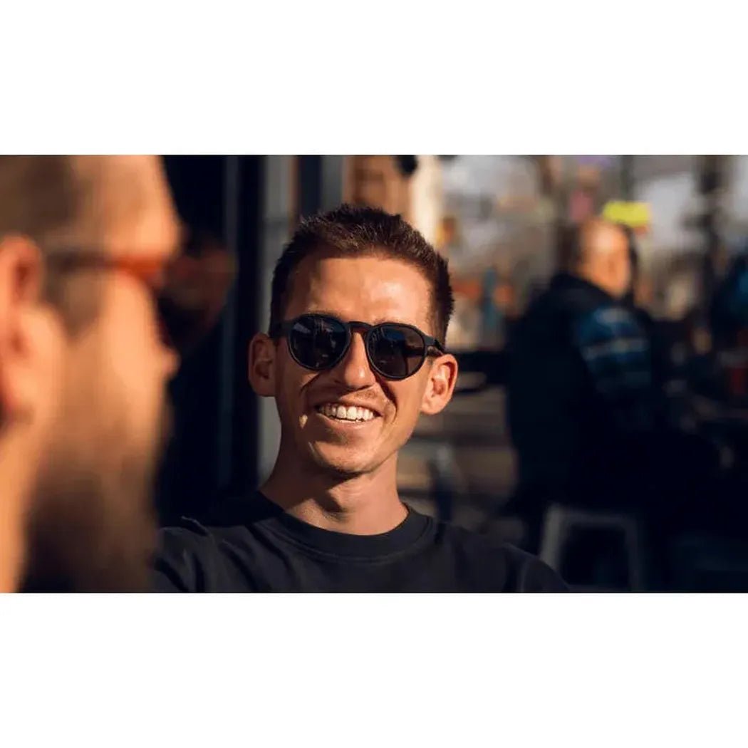 A smiling man wearing Optic Nerve RiNO sunglasses takes a selfie outdoors with a blurred city background.