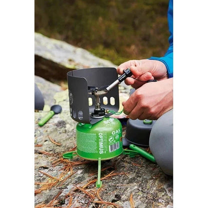 A person lighting the Optimus Sparky camping stove on a rocky outdoor surface with pine needles.