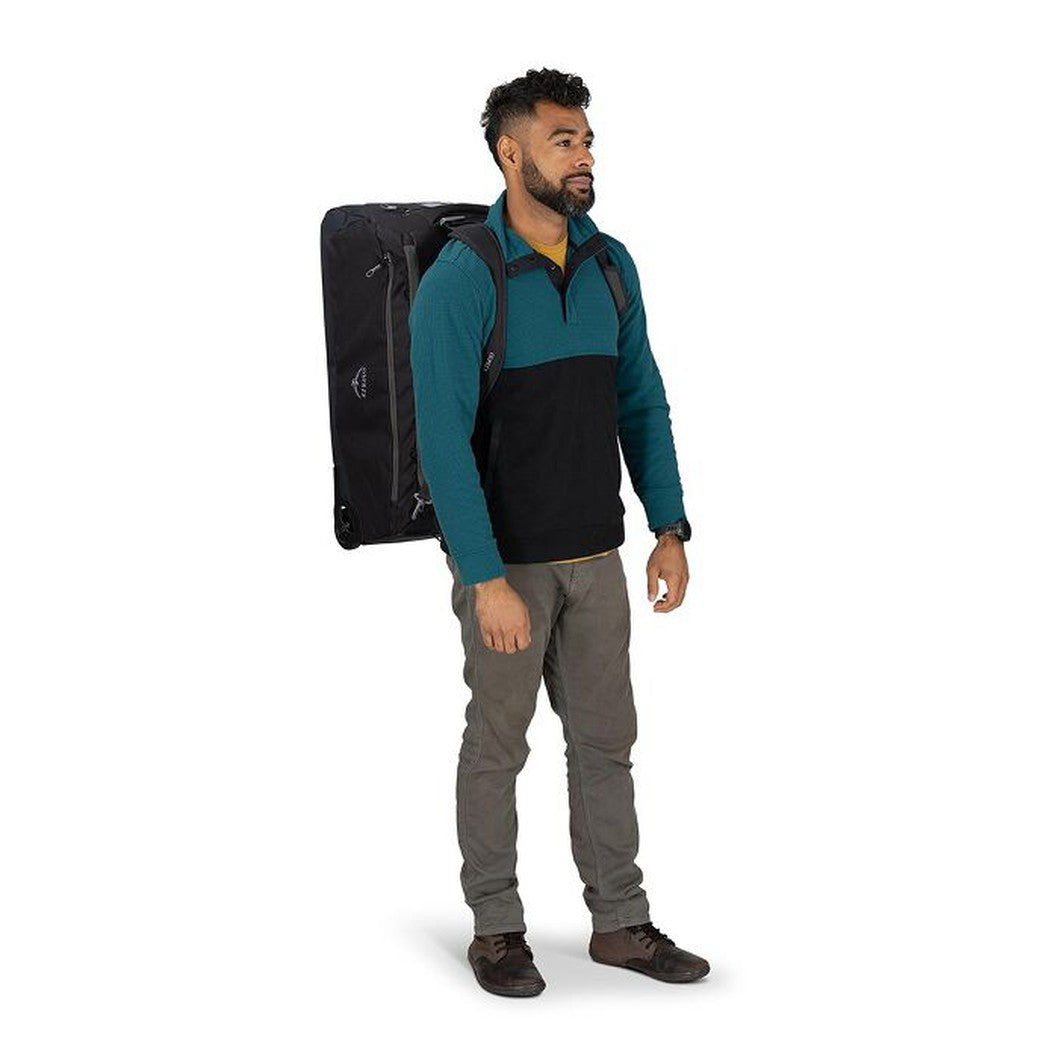 A man stands in a side view wearing the black Osprey Daylite Wheeled Duffel 85 against a white background.