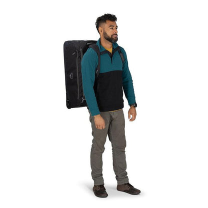 A man stands in a side view wearing the black Osprey Daylite Wheeled Duffel 85 against a white background.
