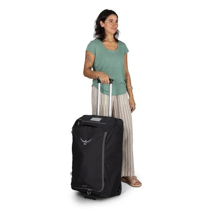 A woman stands beside the black Osprey Daylite Wheeled Duffel 85 on a white background.