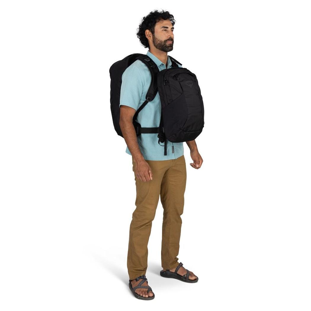 A man wearing a light blue shirt and tan pants stands in a side view with a black Osprey Farpoint 55 backpack against a white background.