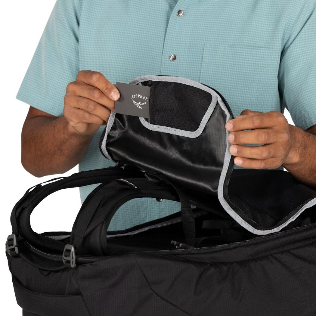 A person in a light blue shirt holds up a tag inside the open black Osprey Farpoint 55 luggage against a white background.