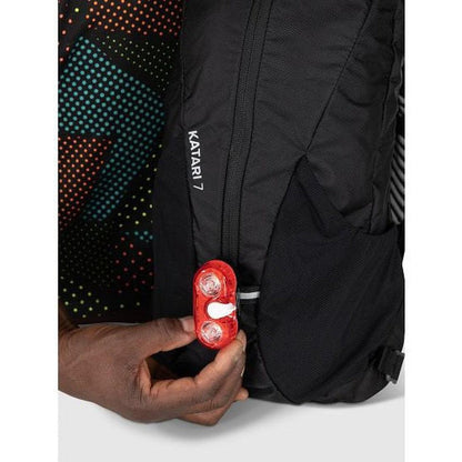 A hand holds a red light near the Osprey Katari 7 black backpack with "Katari 7" text visible.