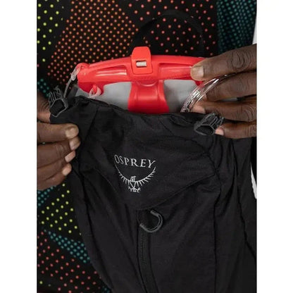 A close-up of the Osprey Katari 7 hydration pack in black, showing the red reservoir and the Osprey logo on the front pocket, with a colorful dotted background.