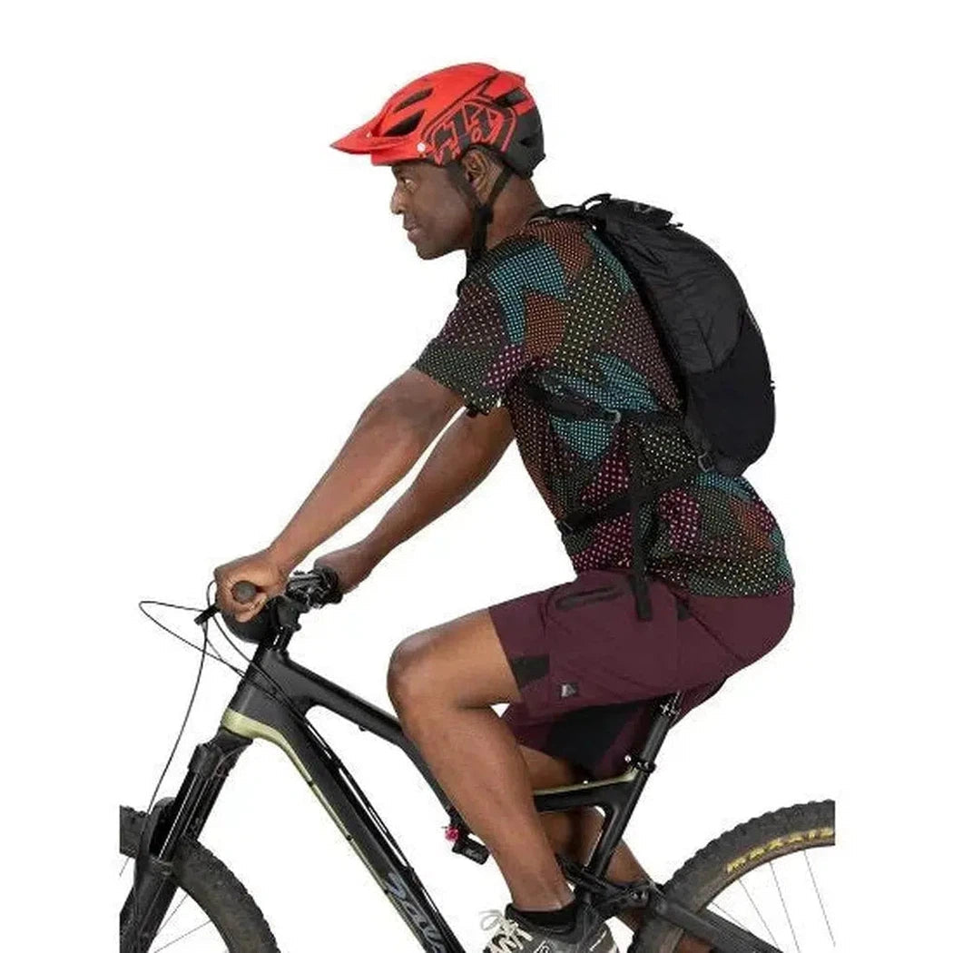 A man wearing a red helmet rides a bike while wearing the black Osprey Katari 7 hydration pack against a white background.