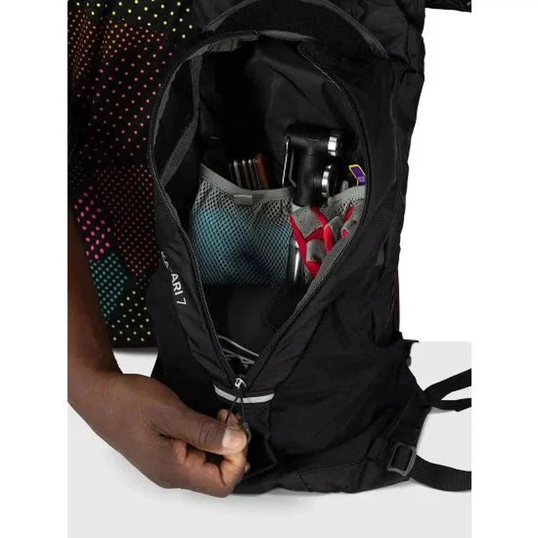 A hand zips open the black Osprey Katari 7 backpack, revealing a mesh pocket with a camera and other gear inside.