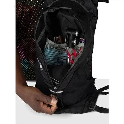 A hand zips open the black Osprey Katari 7 backpack, revealing a mesh pocket with a camera and other gear inside.