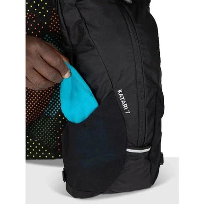 A hand pulls a blue item from the front pocket of the black Osprey Katari 7 backpack, which has a white stripe near the bottom.