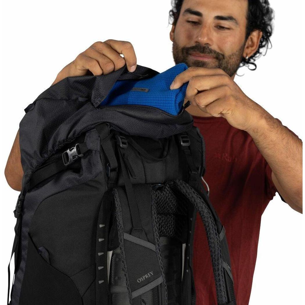 A man in a red shirt puts a blue item into the top pocket of the black Osprey Kestrel LT 65 backpack against a white background.